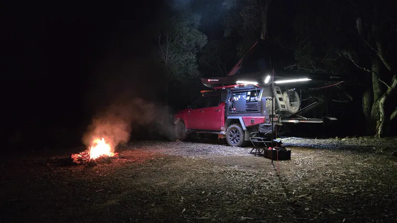 Campfire beside the red Hilux at night, its canopy lights on